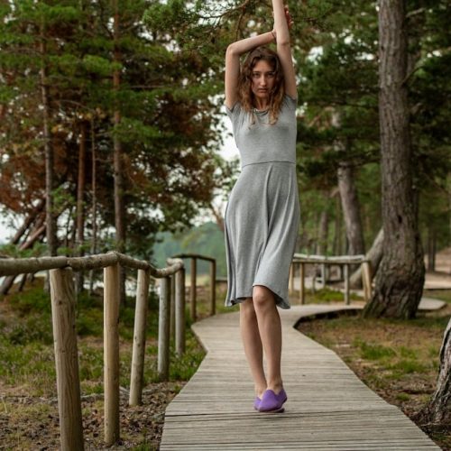 A slim dark-haired woman in a gray dress walking along a wooden boardwalk by the sea, surrounded by coastal forest.