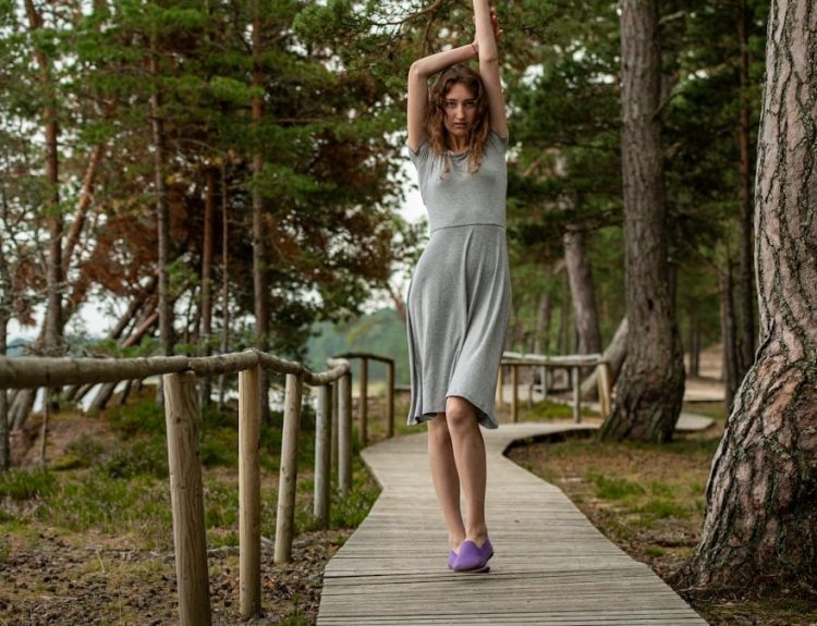 A slim dark-haired woman in a gray dress walking along a wooden boardwalk by the sea, surrounded by coastal forest.